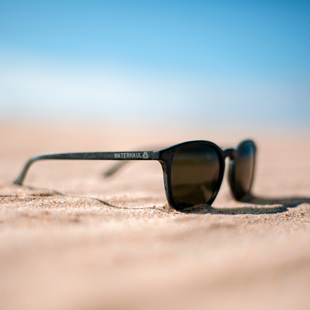 Waterhaul sunglasses with matte dark grey frames and brown polarized lenses, resting on sunlit sand with a blurred ocean and sky backdrop. Sustainable eyewear crafted from recycled ocean plastic, captured in a natural coastal setting.