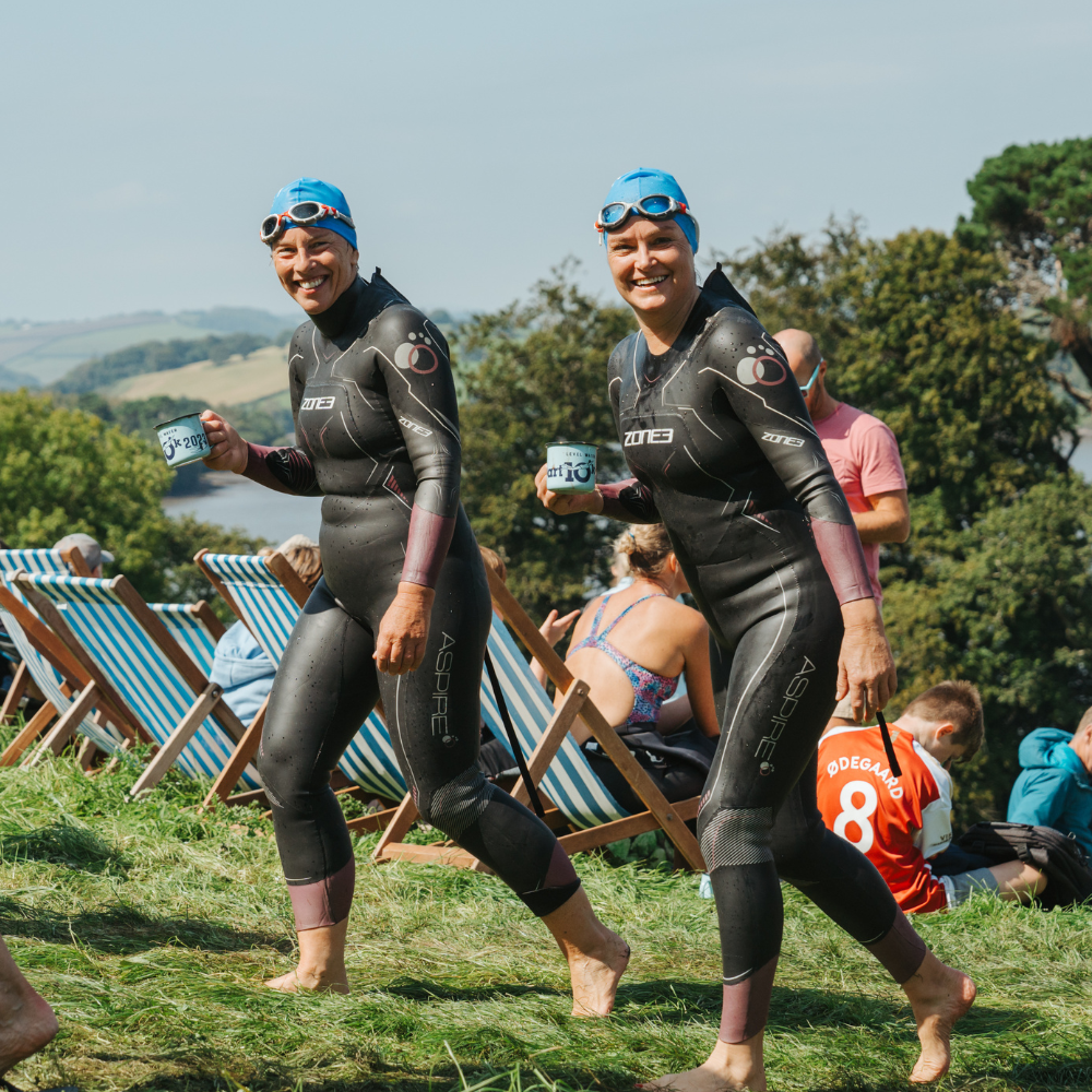 2 women wearing ZONE3 wetsuits. They are smiling at the camera, wearing blue swim caps and holding cups.