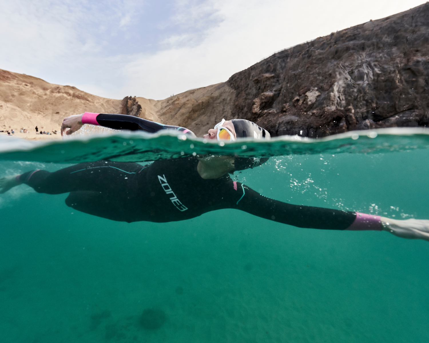 Person swimming in clear blue water in Agile wetsuit and goggles, surrounded by rocky cliffs.
