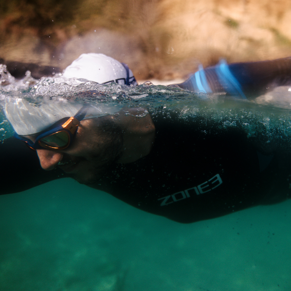 Male swimmer in a Zone3 wetsuit with blue accents, white swim cap, and mirrored goggles, captured mid-stroke at the water’s surface in a split-level underwater shot. Showcasing endurance and performance in a natural open-water environment.