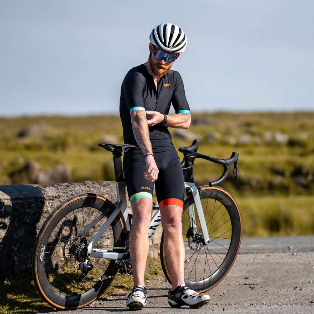 Man in ZONE3 Activate+ trisuit in black standing beside a road bike, adjusting his sleeve while wearing a helmet and sunglasses.