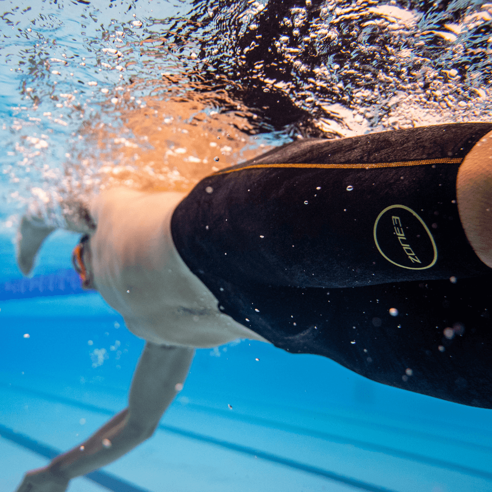 Underwater view of a swimmer in black MF-X jammer with a gold circular logo moving through the pool
