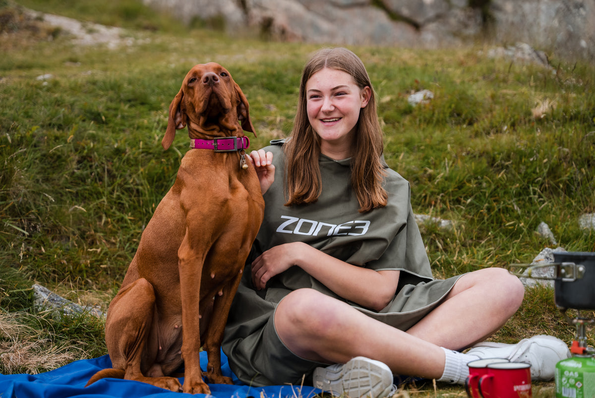 A girl wearing the Khaki Microfibre Changing Robe sat with a  dog by the lake.