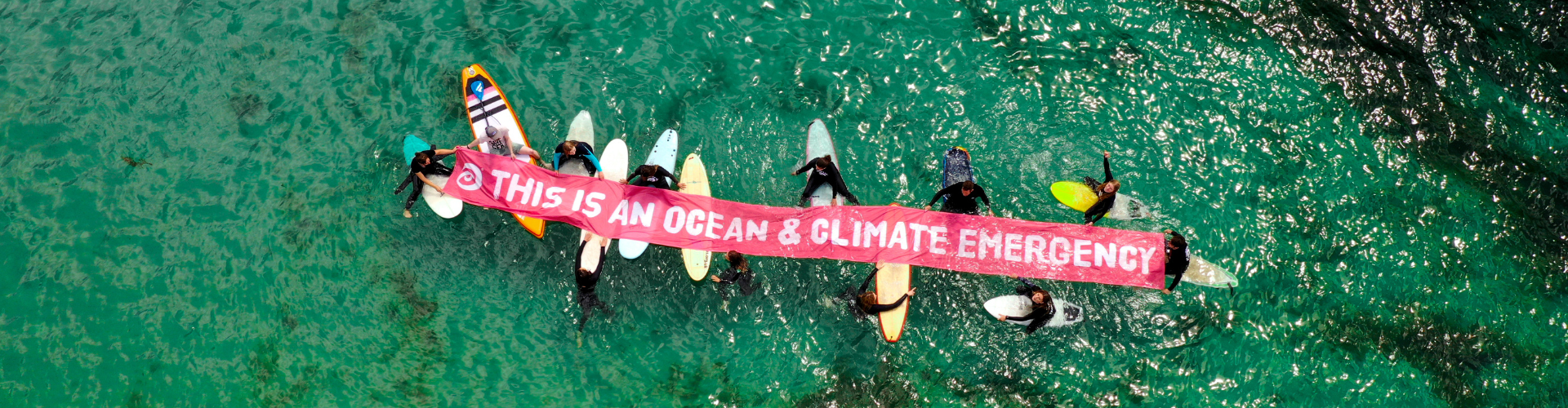 Top down image of surfers in the water. They are holding a banner that reads "This is an ocean and climate emergency"