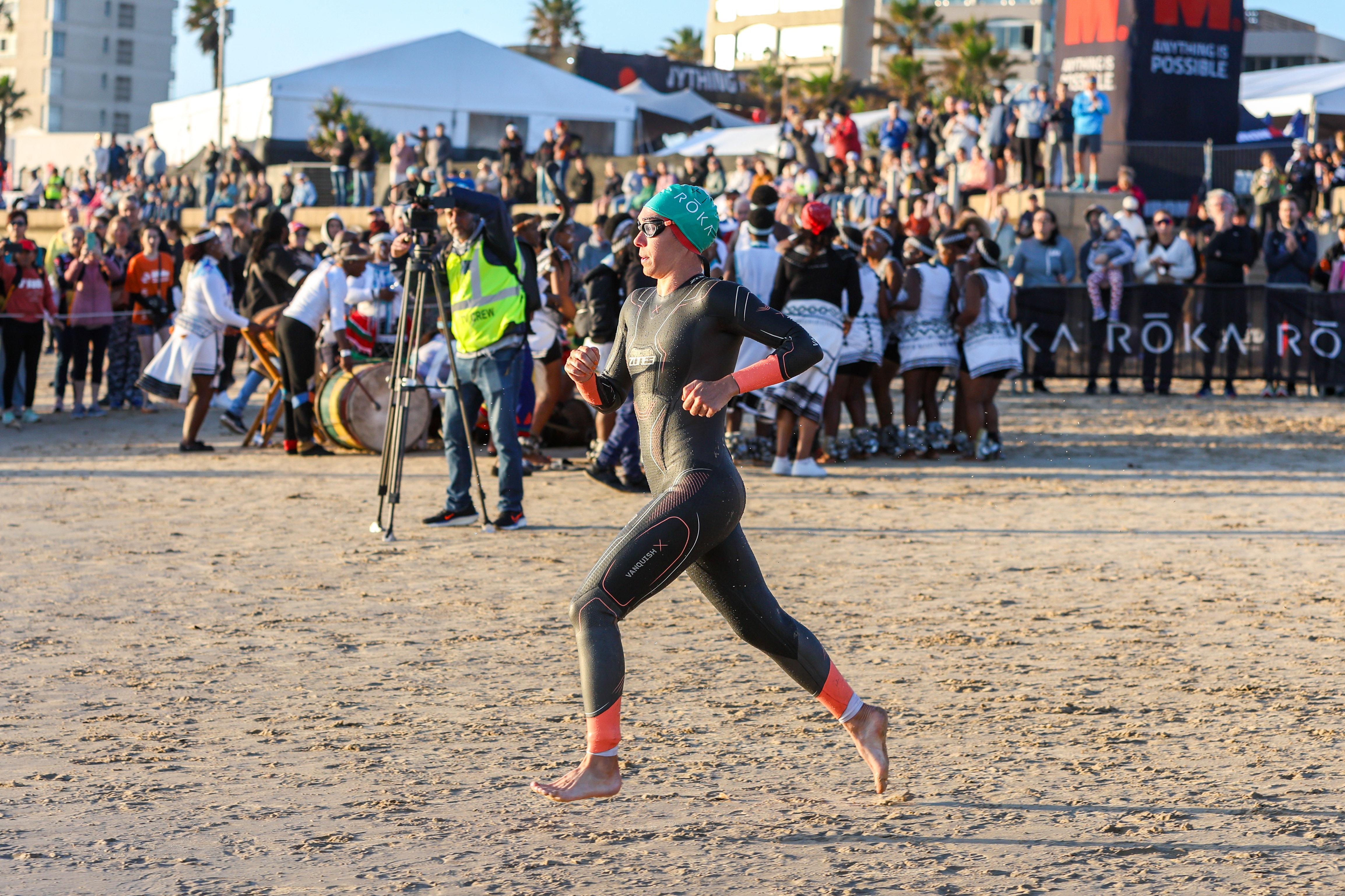 Person in a wetsuit running on a beach with a crowd in the background