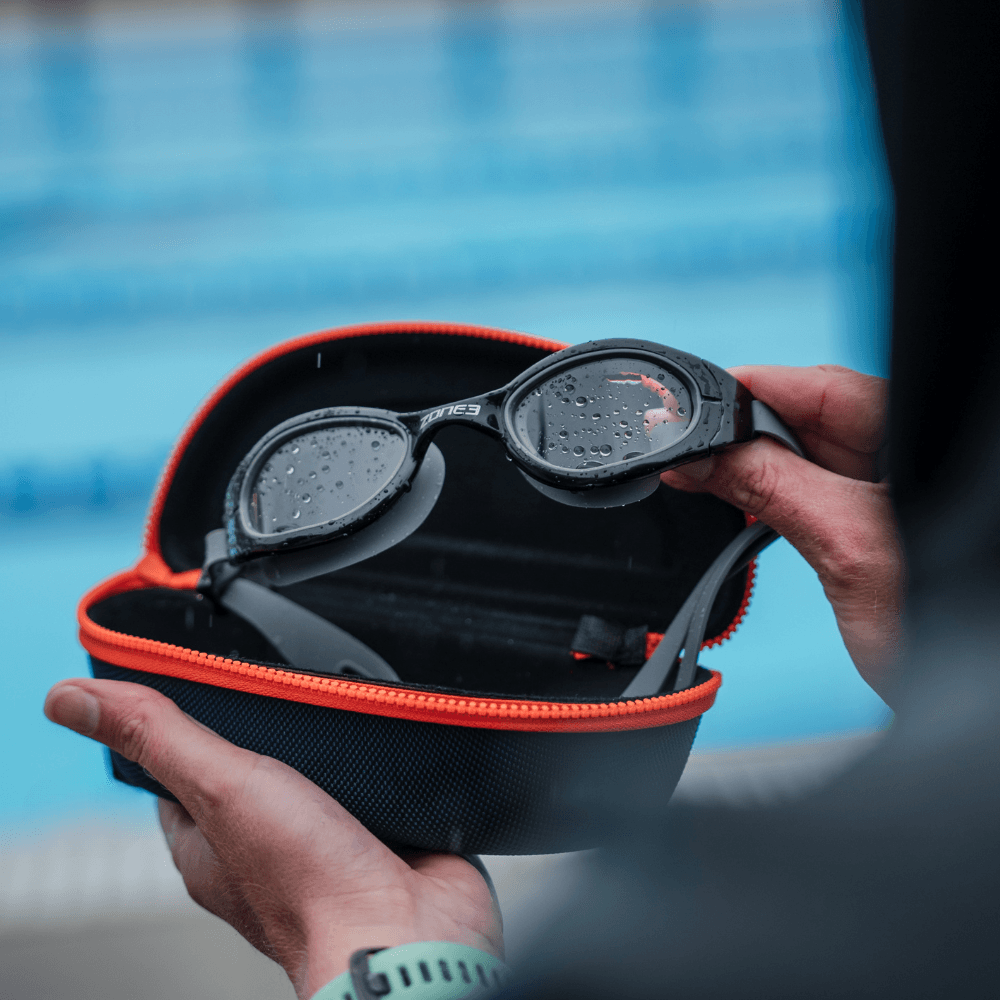 opened Navy blue and orange case with Zone3 logo. showing someone taking out their Zone3 Goggles out the protective case. Blurred pool background.