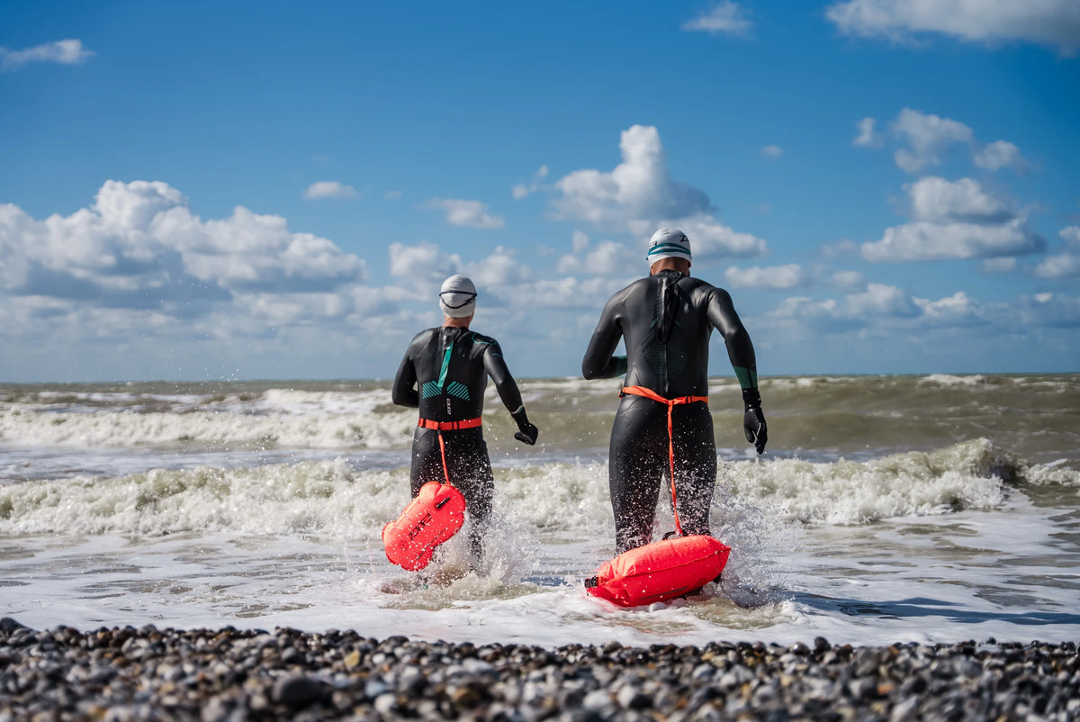 Two swimmers running into open water fully prepared with Zone3 wetsuits and the Recycled Safety Buoy Dry Bag 28L for visibility.