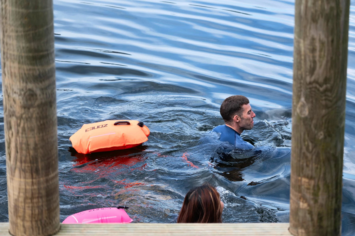 Two swimmers swimming in open water with an orange and pink recycled safety buoy. 