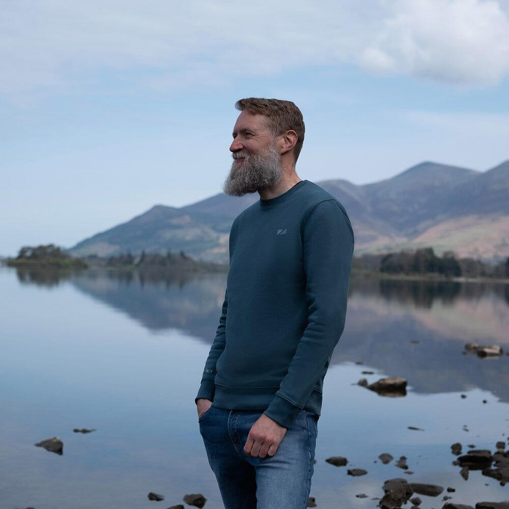 A Man standing by a lake wearing the Zone3 Seathwaite Crew Neck Sweatshirt in blue paired with jeans. 