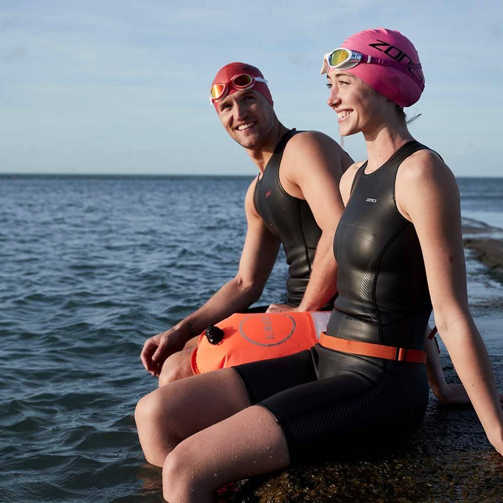 Athletes wearing the Zone3 swim cap, goggles sitting on a rock at the beach. 