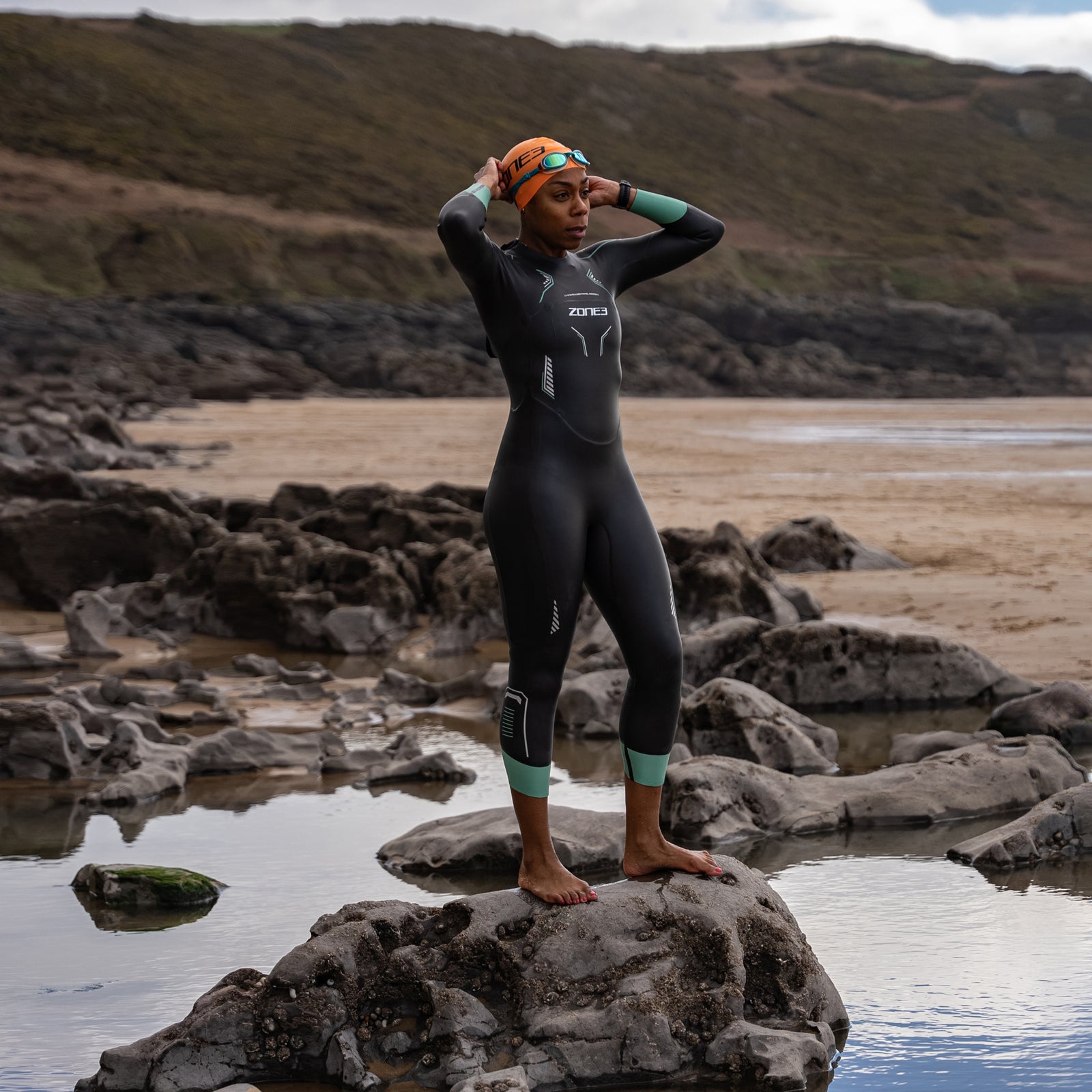 A lady standing on a rock wearing a ZONE3 vision wetsuit. She is also wearing an orange ZONE3 swim cap and some teal ZONE3 goggles. The background is rocky and sandy.