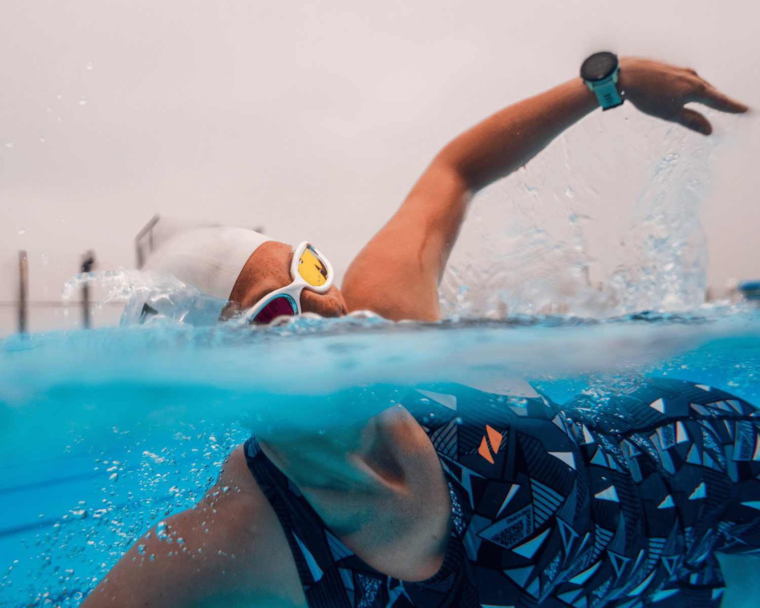 A swimmer in a patterned swimsuit and white swim cap performing a freestyle stroke in an outdoor pool, with water splashing around them and an overcast sky in the background.