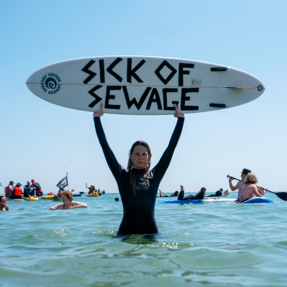 An image of a lady in a wetsuit. She is standing in the sea holding a surf board that reads "Sick of sewage".