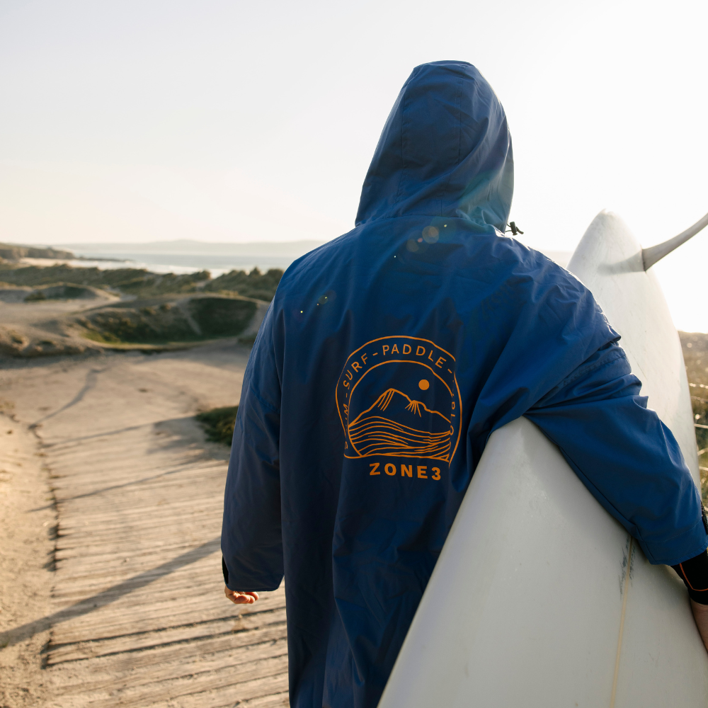 Person in a blue ZONE3 robe with hood up, walking with a surfboard under arm, large logo on back visible.