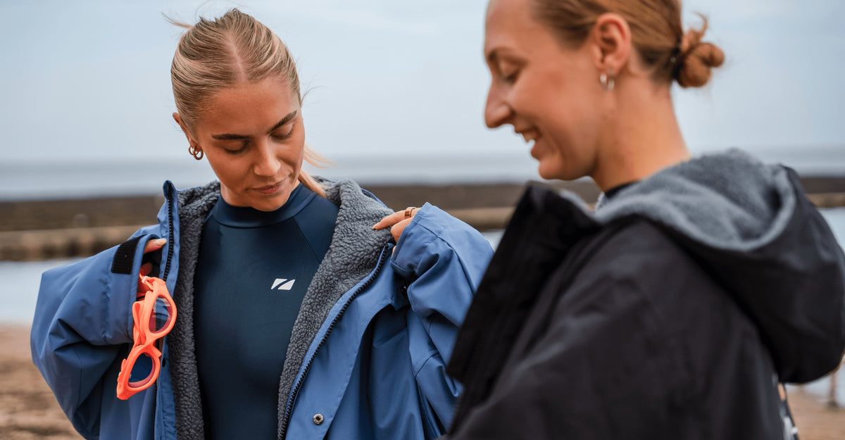 Women wearing the Zone3 Ti+ long sleeve swimsuit with the Zone3 rob on top at the beach. 