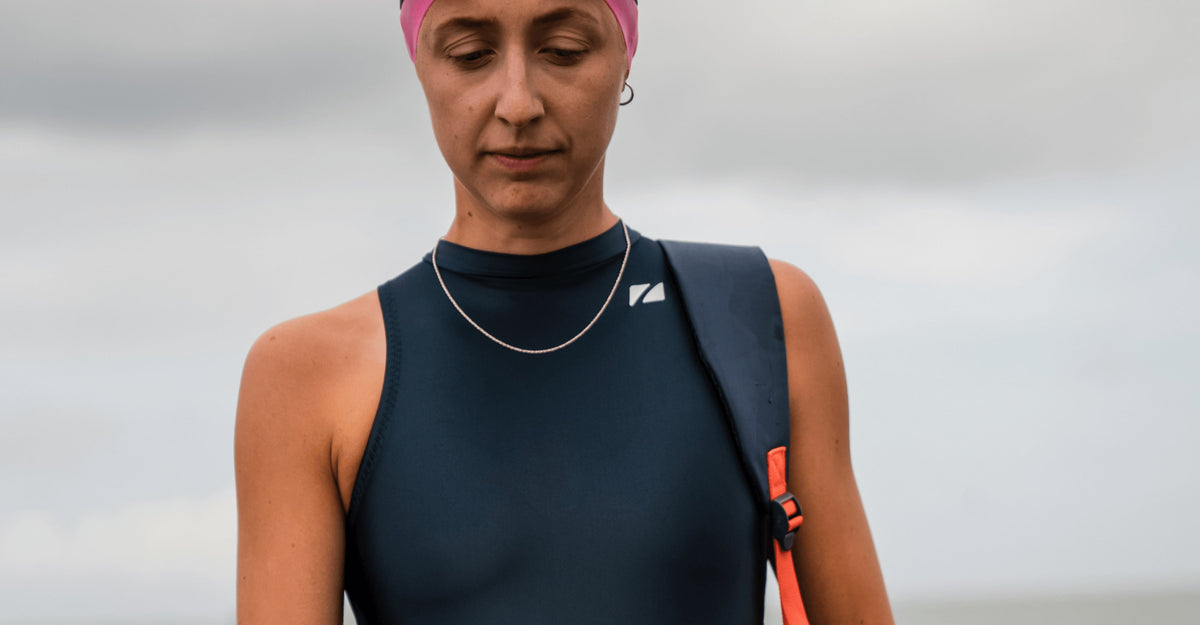 close up of A woman wearing the Zone3 Deep sea Ti+ Thermal High Neck Swimsuit. one-piece swimsuit, a pink Zone3 swim cap, standing on a beach with a cloudy sky in the background.