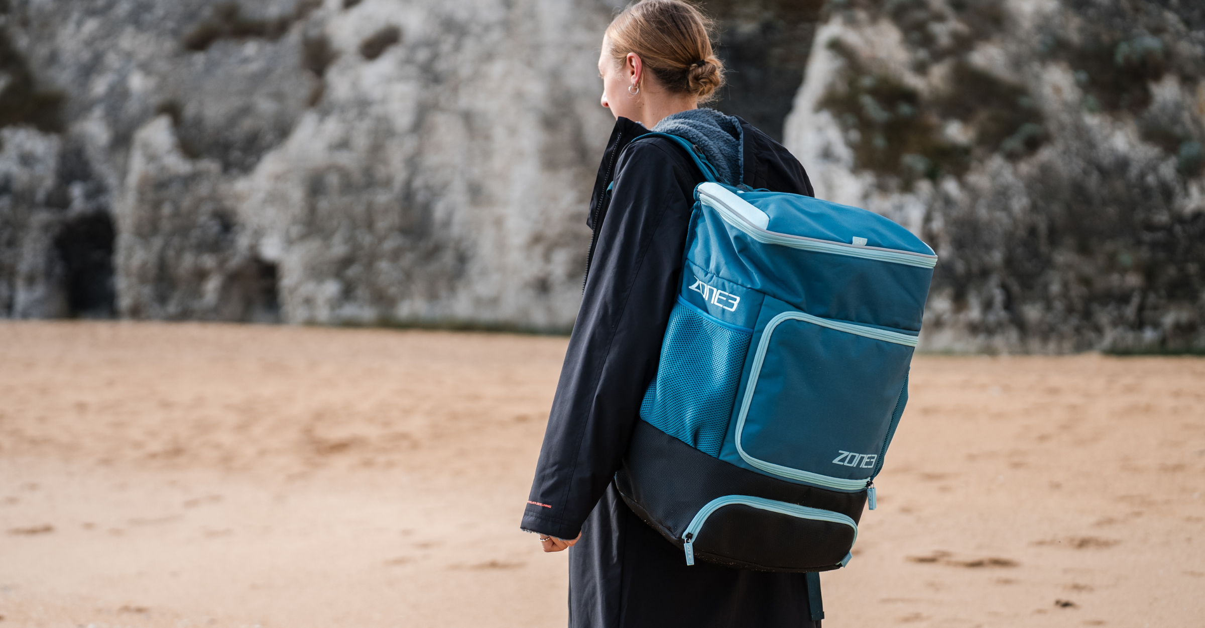 Person with Zone3 teal transition backpack standing on a beach with a rocky cliff in the background