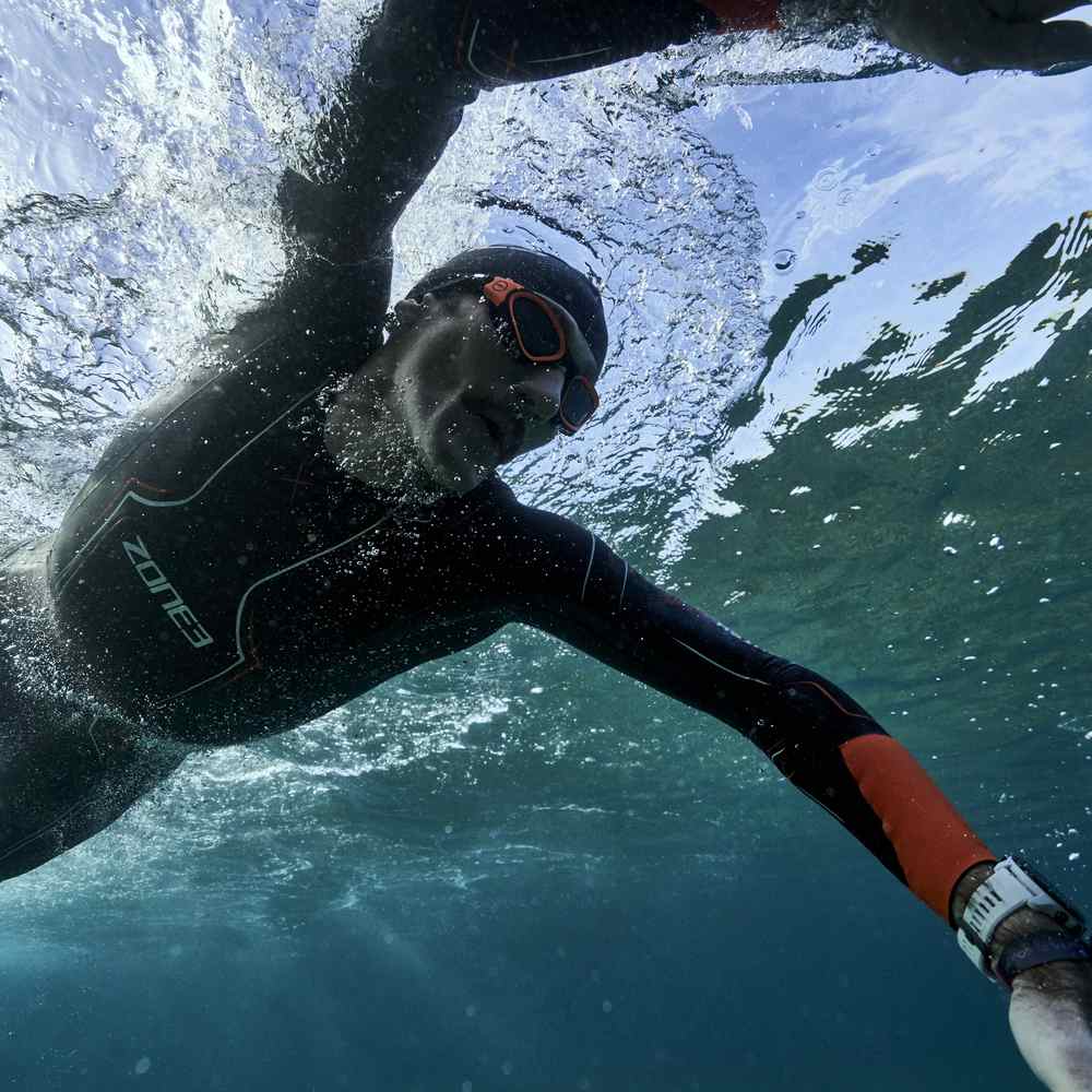A male swimming with his head underwater. The shot is taken from beneath him. He is wearing a ZONE3 Vanquish-X wetsuit, a black ZONE3 swim cap, and orange ZONE3 swim goggles.