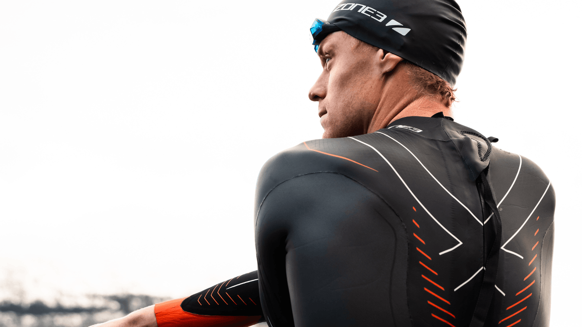 Casper Stornes wearing a black vanquish x wetsuit with red accents, standing outdoors with mountains in the background.