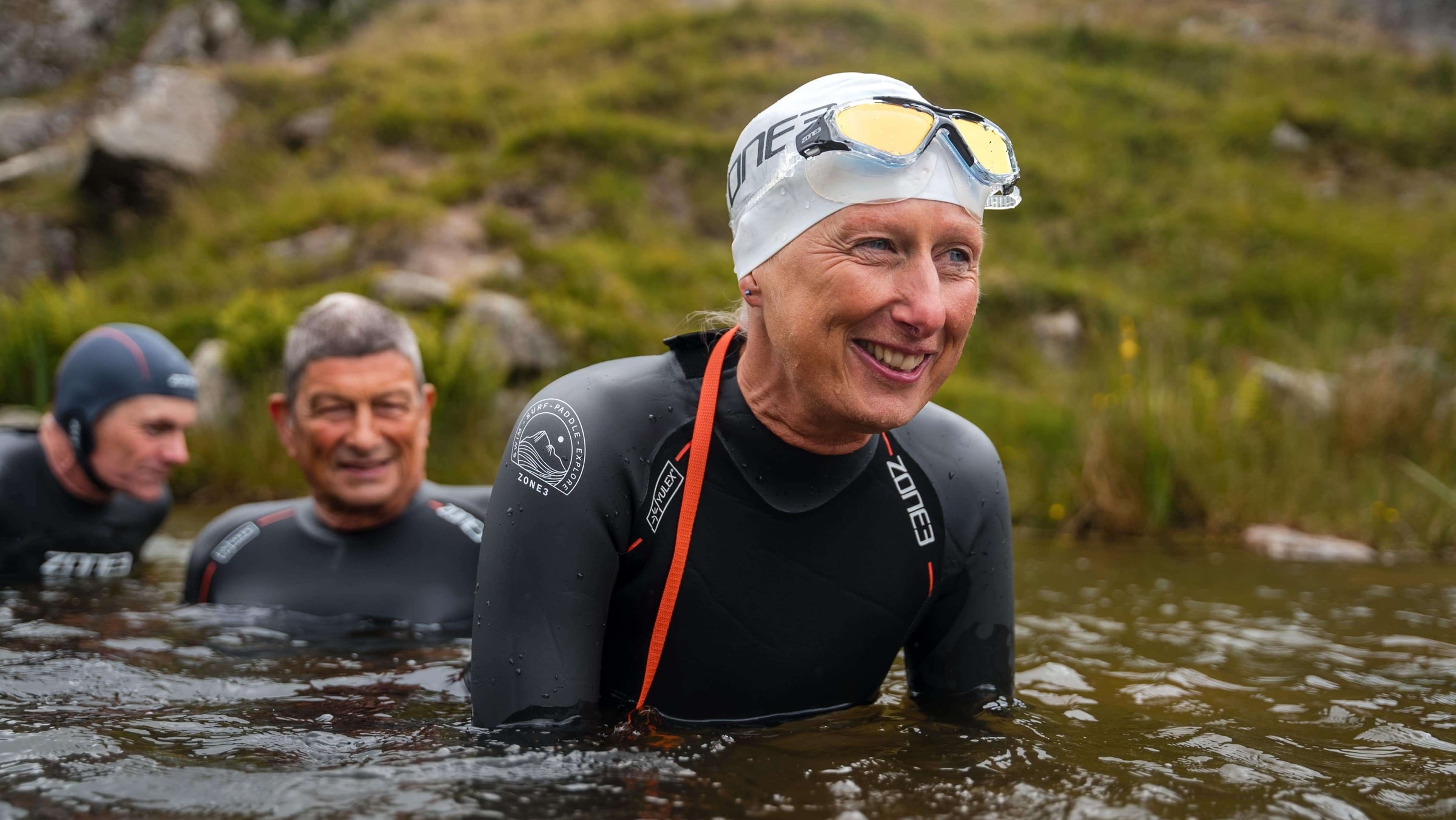 Women in a Zone3 Vision Max Mirror Swim Mask, wetsuit and swim cap emerging from water with two others in the background. 