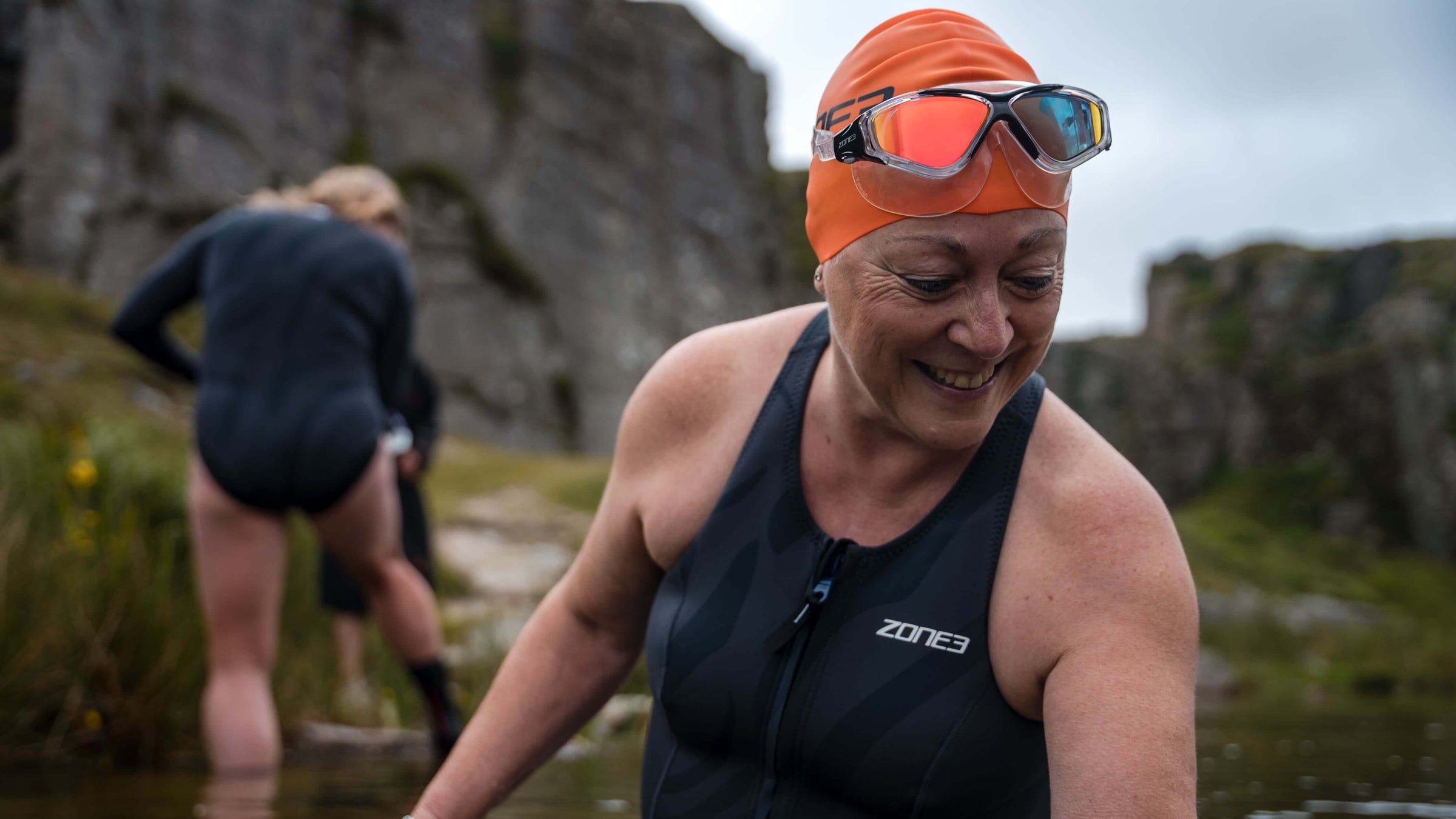 Woman wearing the Zone3 Vision Max Mirror Swim Mask  swim gear and orange swim cap, standing in a natural water body with rocky landscape.