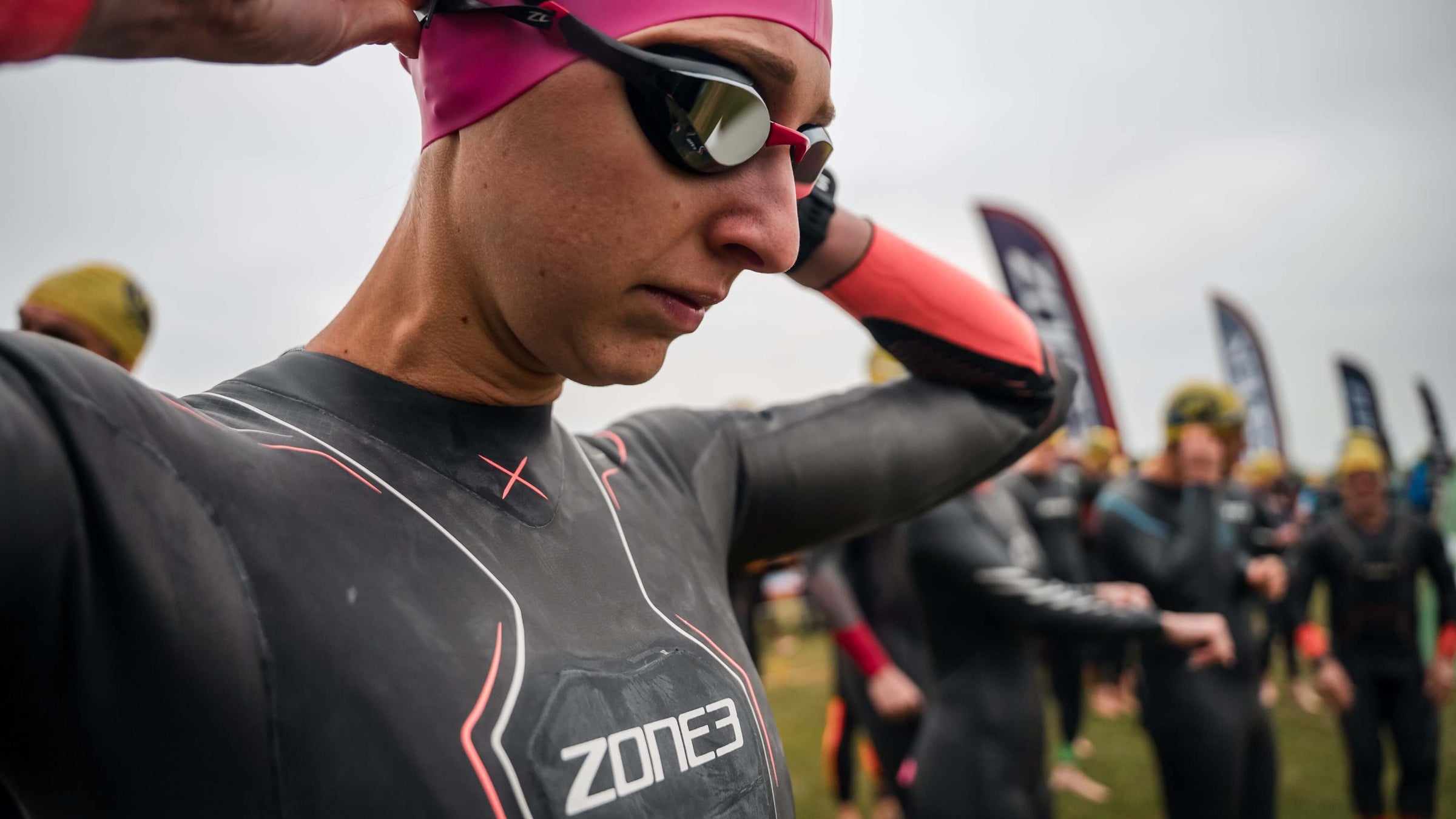 Athlete wearing a Zone3 wetsuit, pink swim cap with  Volare racing swim goggles, preparing for a swim event.