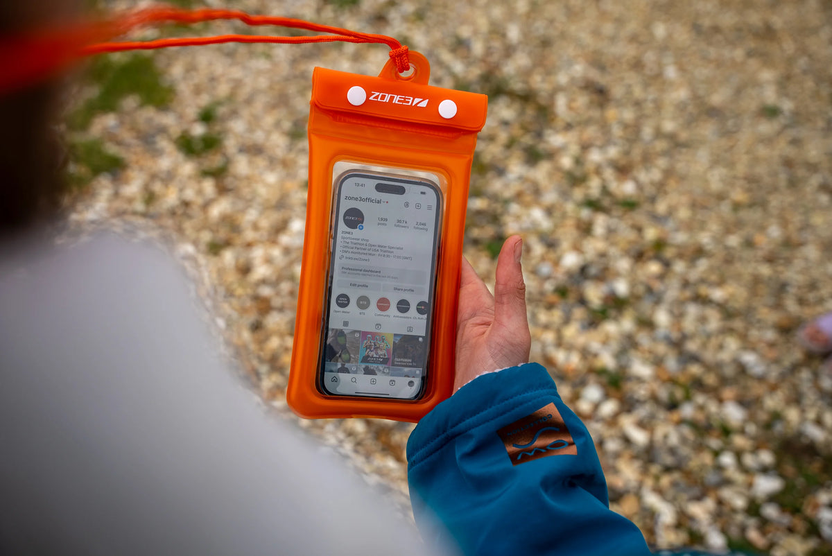 Athlete holding the Zone3 Orange waterproof phone pouch at the beach. 