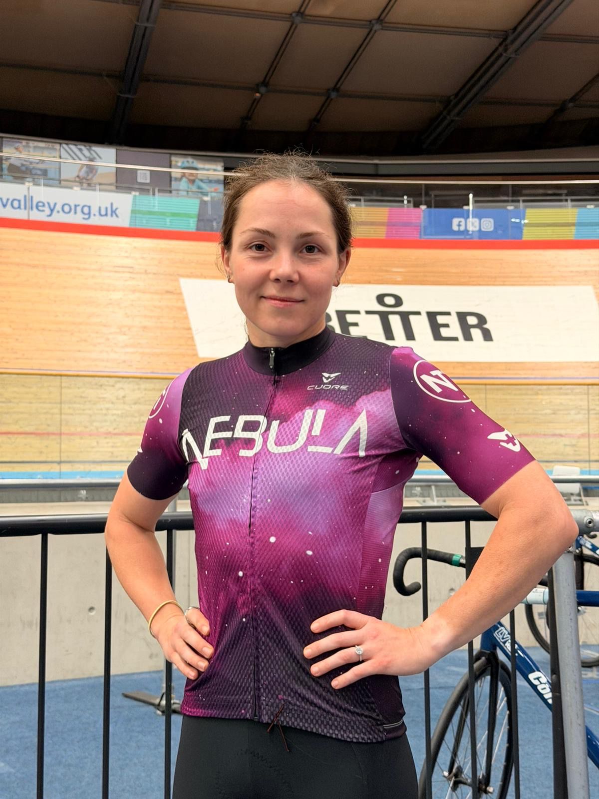 Person wearing a purple cycling jersey with 'Nebula' branding in an indoor cycling track setting.