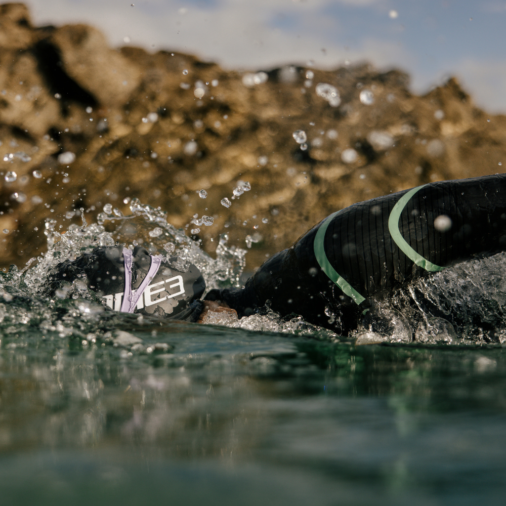 Close-up of a swimmer wearing a Zone3 wetsuit, swim cap, and goggles mid-stroke in open water, with water droplets suspended in motion. Captured from the waterline with a rugged coastal backdrop, showcasing endurance and performance in natural conditions.