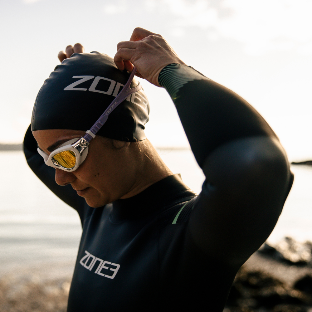 Female swimmer adjusting her Zone3 goggles and swim cap at the water’s edge, wearing a full-length Zone3 wetsuit with green detailing. Captured during golden hour, showcasing pre-swim focus and performance-ready preparation.