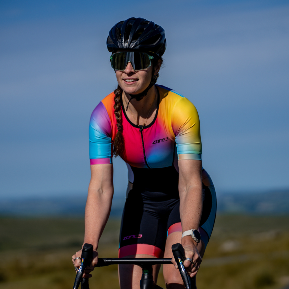 Close-up front view of a cyclist wearing the ZONE3 Activate+ women’s short sleeve trisuit. Gradient colours in blue, pink, yellow, and orange are visible under sunlight.