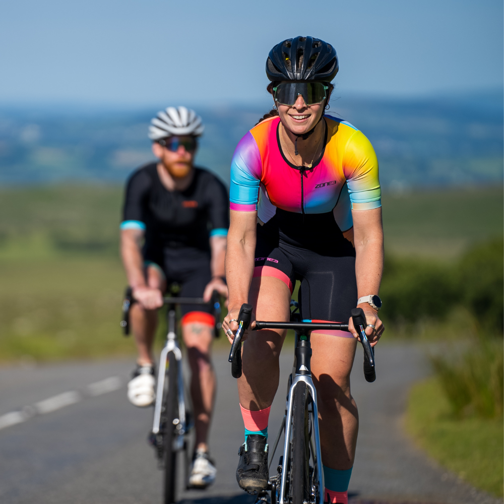 Woman cycling outdoors wearing the ZONE3 Activate+ women’s short sleeve trisuit with a black helmet and sunglasses. Gradient design visible across shoulders and arms.