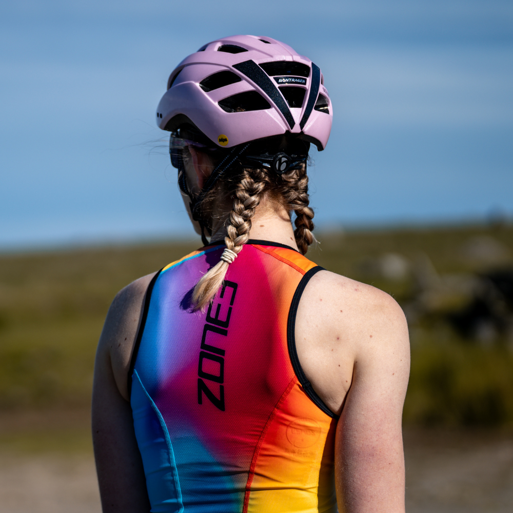 Cyclist wearing the ZONE3 Activate+ sleeveless trisuit with a pink helmet. The trisuit has a vertical ZONE3 logo and a bright gradient design on the back.