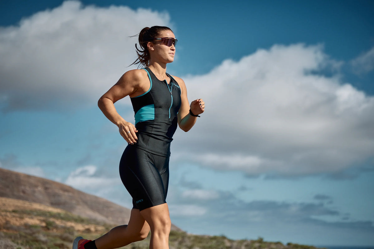Woman running outdoors with a scenic background wearing the Zone3 Activate Tri Top in black and teal.
