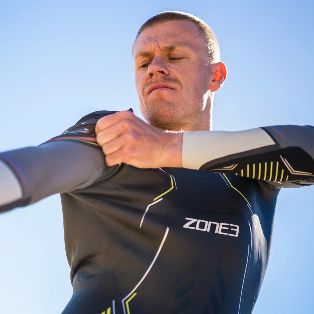 A man outdoors wearing a dark gray and black Zone3 athletic wetsuit, stretching his arm over his shoulder against a clear blue sky.