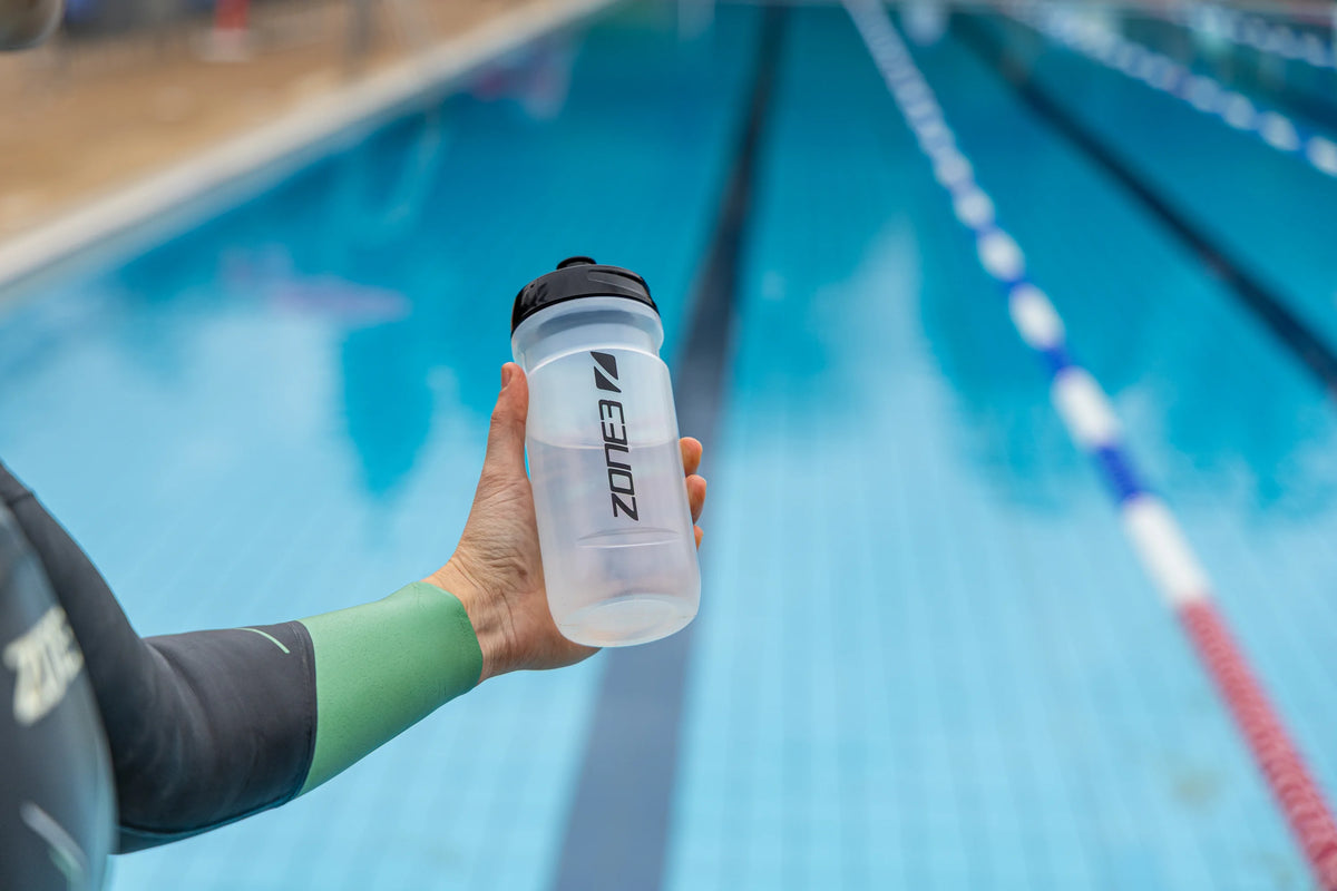 athlete holding the Zone3 transparent water bottle with black detailing. blurred pool background. 