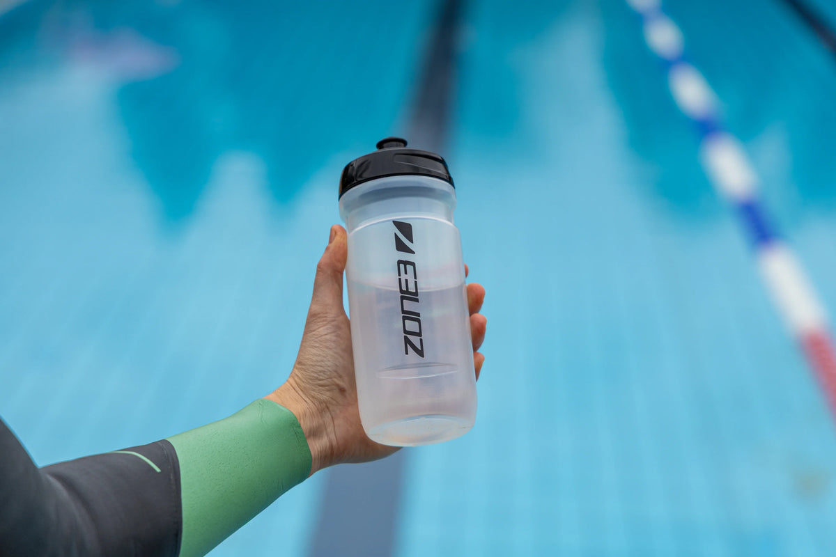 close up of an athlete holding the Zone3 transparent water bottle with black detailing. blurred pool background. 