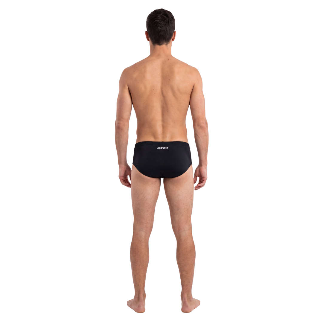 A man viewed from behind wearing black swim briefs with a small white logo on the back waistband, standing against a plain white background.