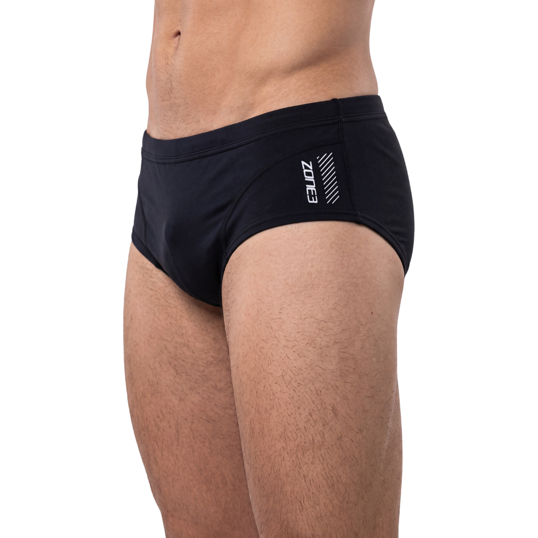 A close-up side view of a man wearing black swim briefs with a white ZONE logo on the hip, set against a plain white background.
