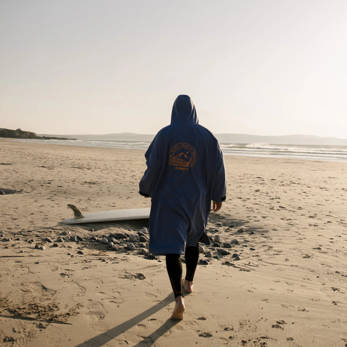 A person wearing a dark blue hooded beach poncho walks barefoot on a sandy beach, with a surfboard lying nearby and the ocean in the distance under a pale sky.