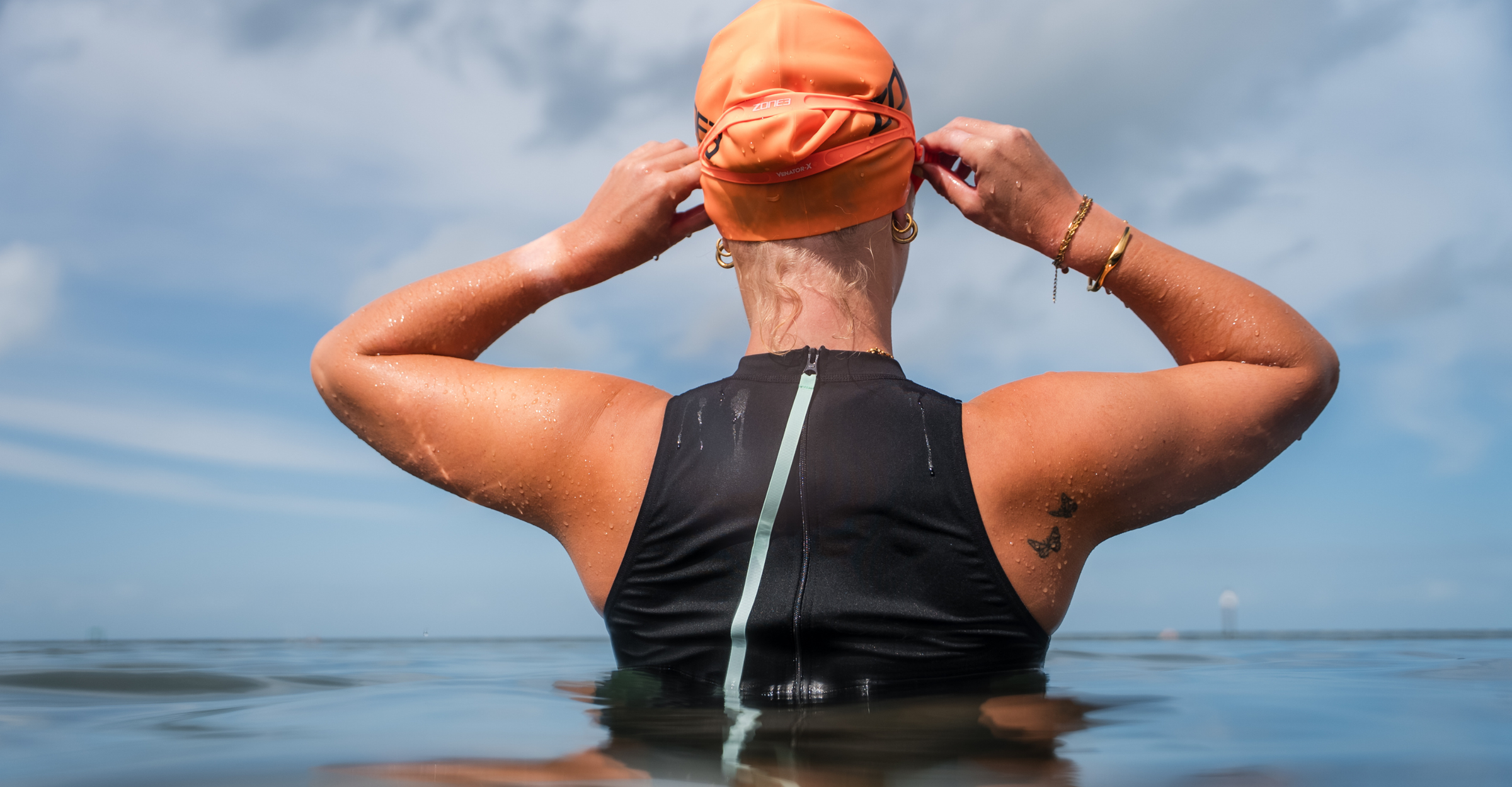 Person wearing an orange swim cap and black high neck swimsuit in water with a blue sky background