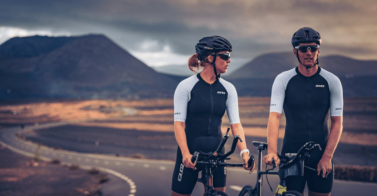 Two cyclists wearing matching Zone3 Lava trisuit with mountains in the background.