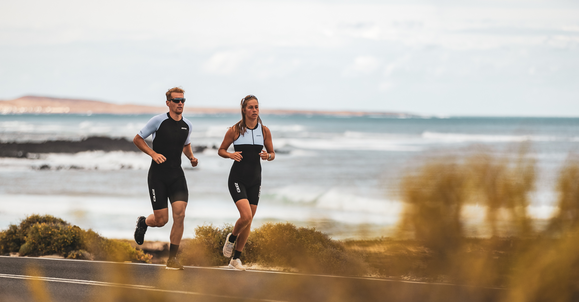 Two runners in lava trisuit running along a coastal road with ocean waves in the background.