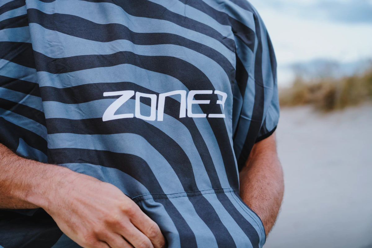 Close-up of a person wearing a short-sleeve microfibre changing robe with horizontal wavy stripes in slate blue and dark gray, featuring the white 'ZONE3' logo on the chest. The background is a blurred outdoor beach or dune setting.