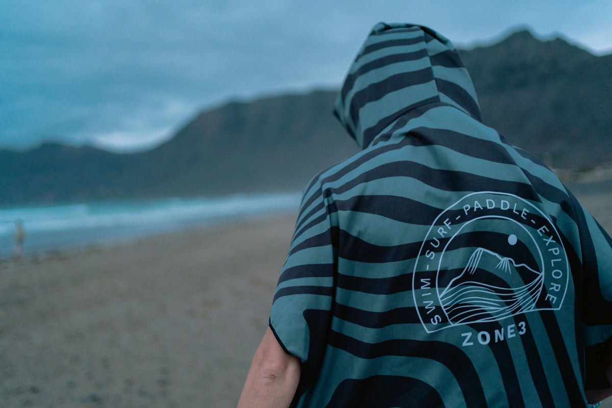 A person standing on a beach wearing a striped changing robe with a white 'Surf Paddle Explore ZONE3' logo on the back, with mountains and ocean in the background.