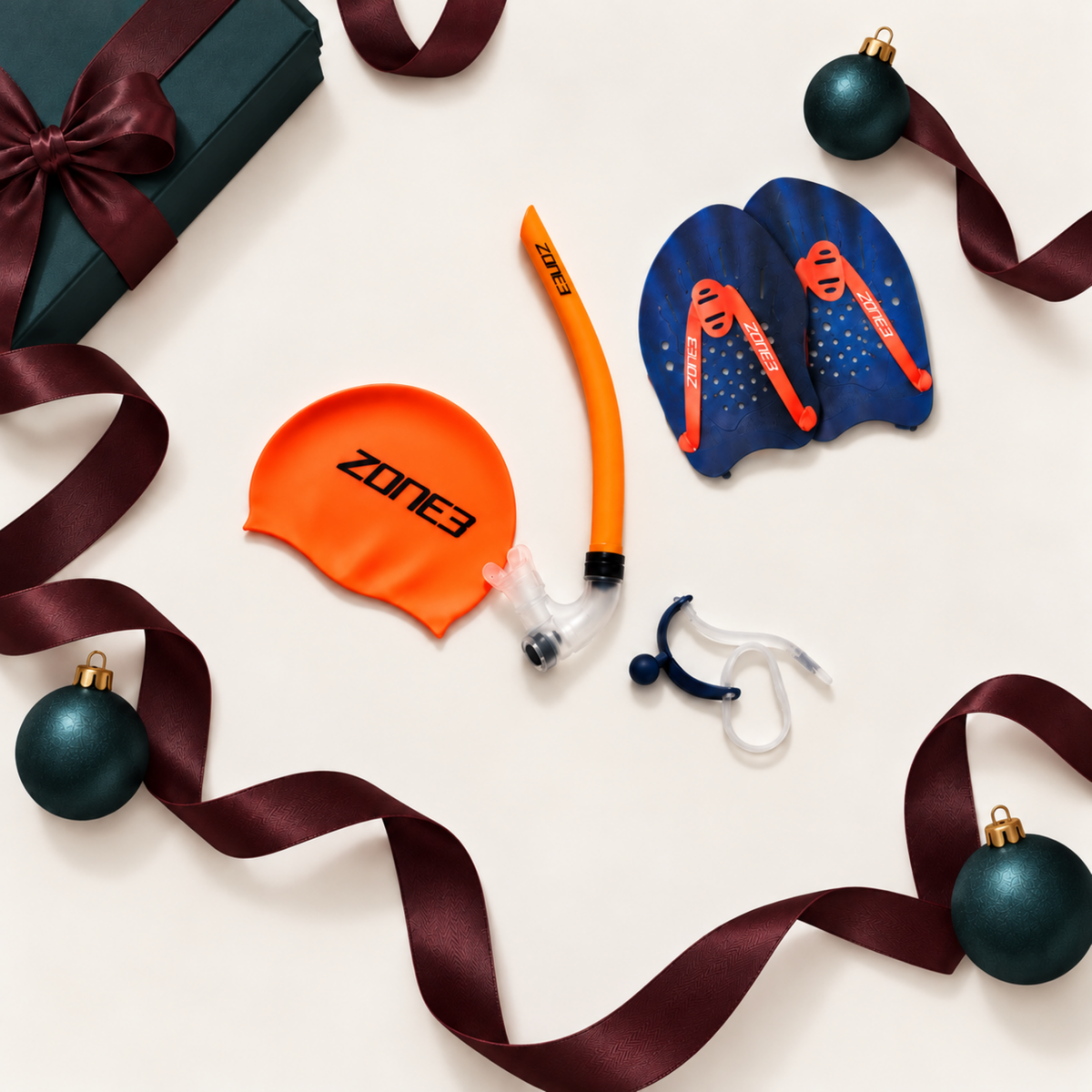 A swimming accessory set laid out on a white background, including an orange snorkel with a clear mouthpiece, two dark blue swim goggles with orange straps, and an orange swim cap with the brand name ZONE3 printed in black.