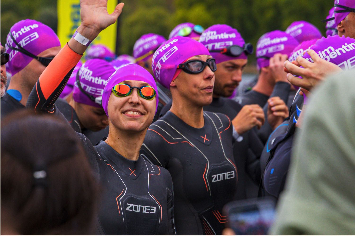 A group of athletes wearing wetsuits and purple swim caps. The two athletes in focus are wearing ZONE3 Vanquish-X wetsuits. One of the two athletes is waving at the camera. 