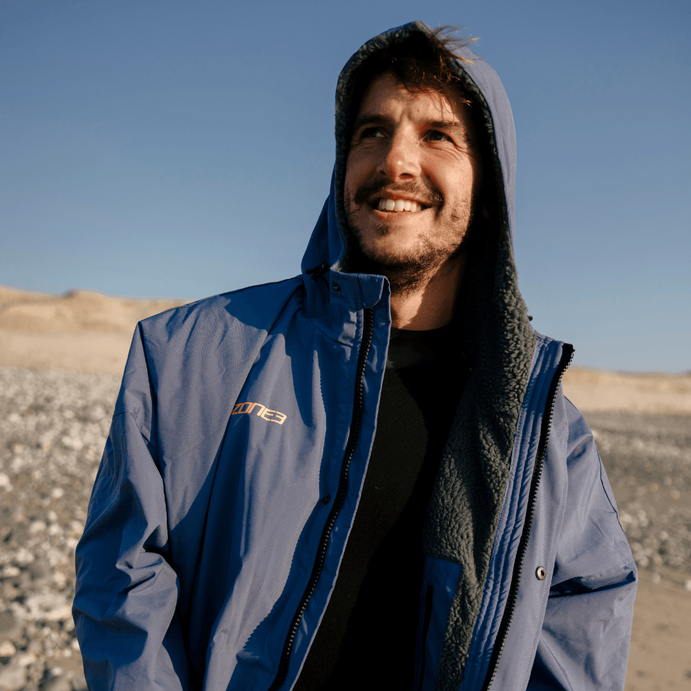 Athlete wearing the blue Thermotech Changing Robe from the front with the hood up at the beach. 