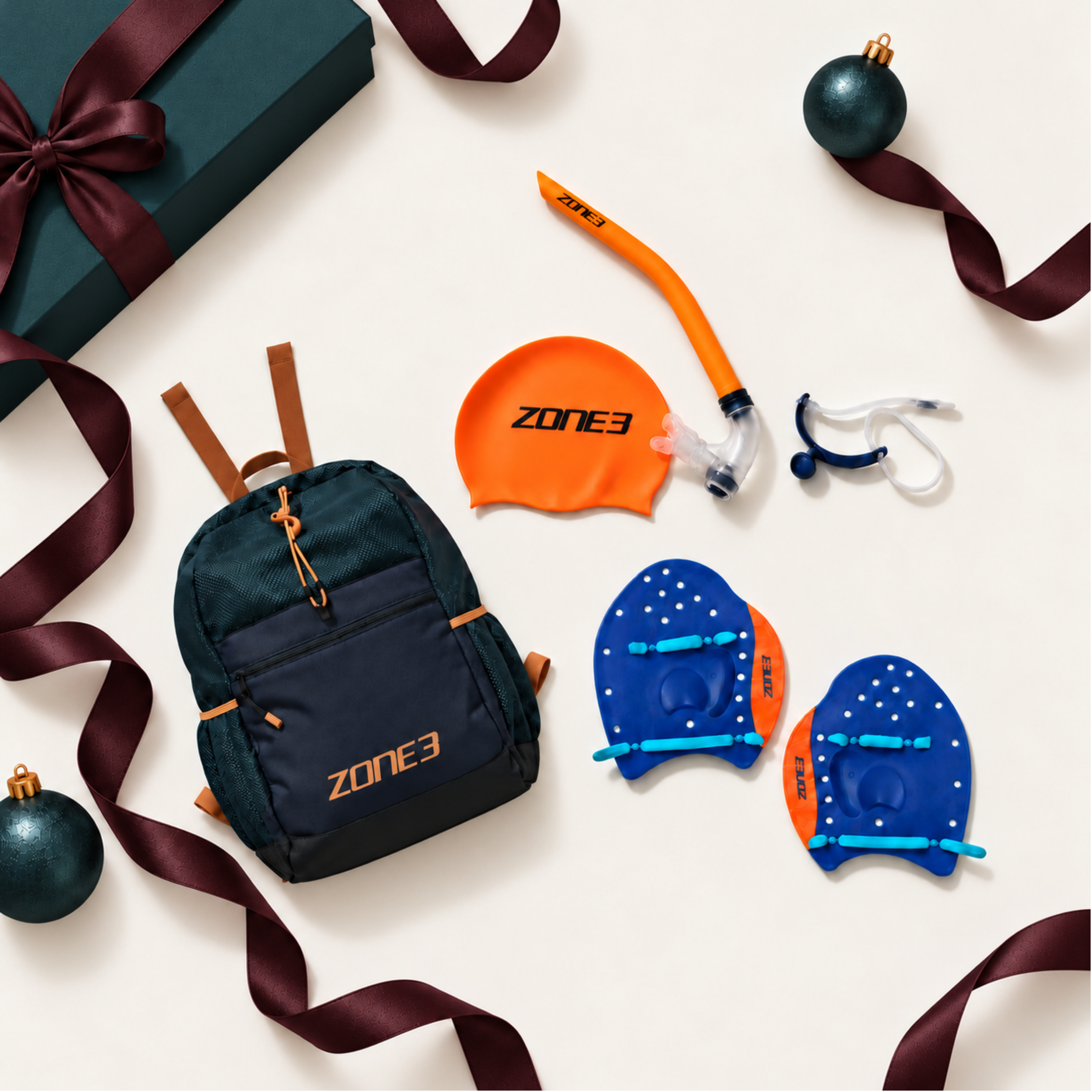 A Zone 3 swim training kit including a navy blue drawstring backpack with orange accents, an orange swim cap, a blue and orange mouthguard, and an orange snorkel mask, arranged on a light gray studio background.