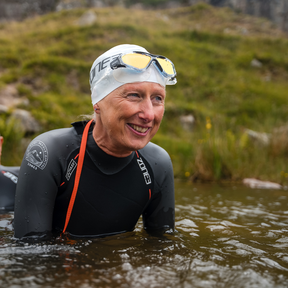 Lächelnde Schwimmerin mit weißer Zone3-Badekappe und Vision Max-Schwimmmaske auf der Stirn, teilweise im Wasser im Freien eingetaucht.